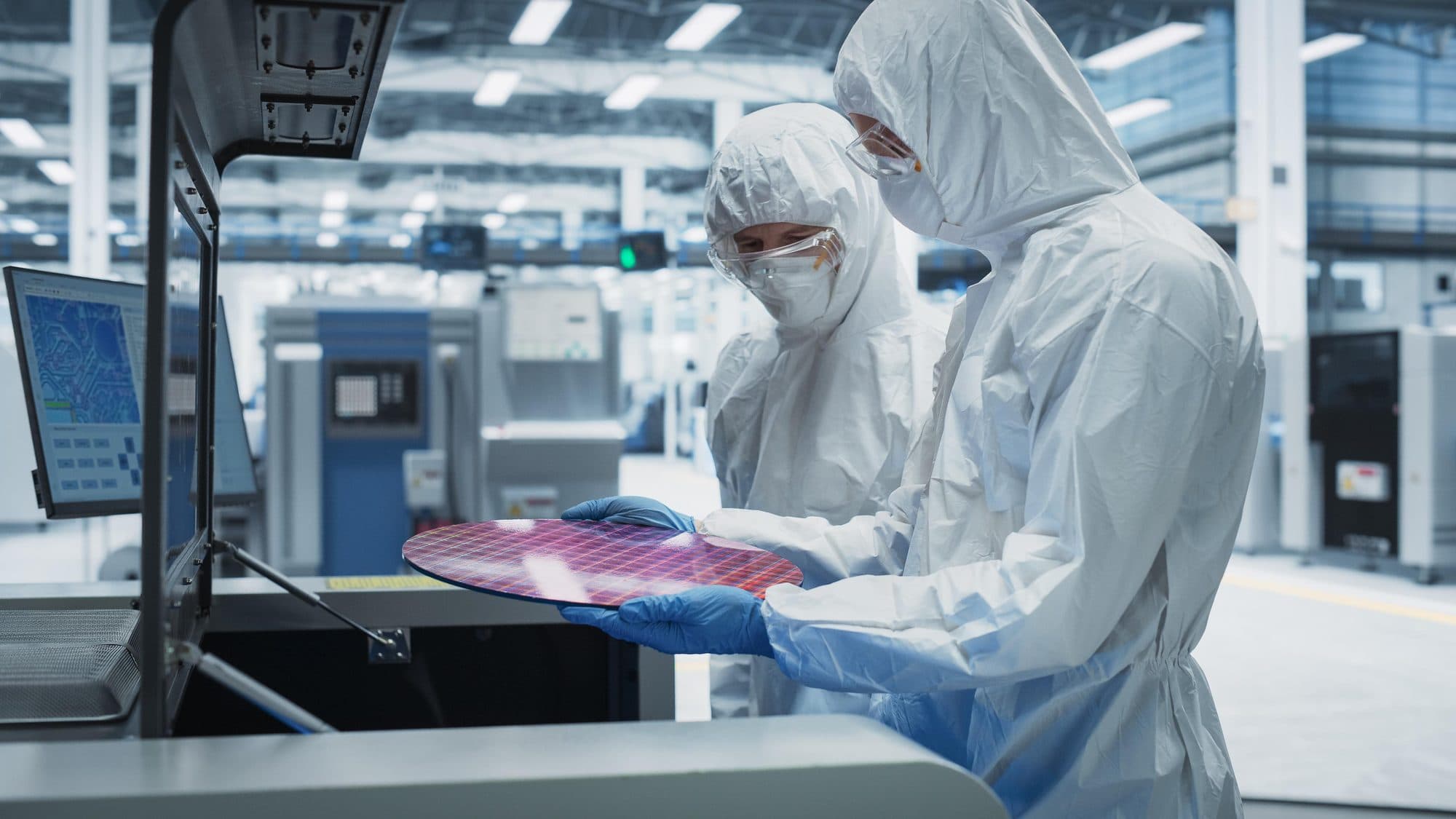 Two engineers in cleanroom suits inspecting a semiconductor wafer on a fabrication line.